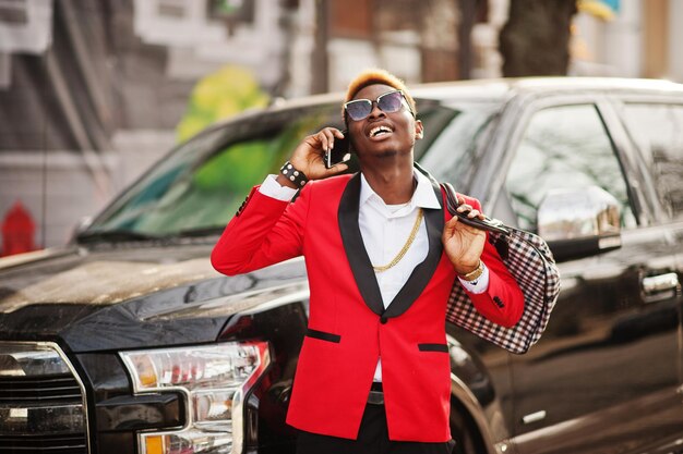 Modelo de hombre afroamericano de moda en traje rojo con reflejos en el cabello posado contra un gran automóvil negro y hablando por teléfono móvil