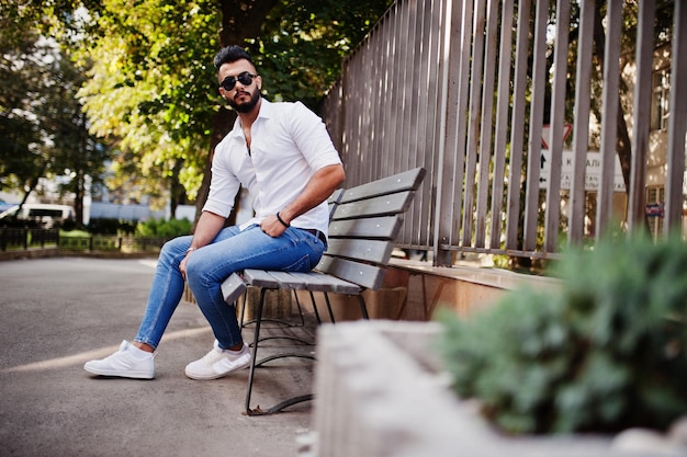 Modelo de hombre árabe alto con estilo en pantalones vaqueros de camisa blanca y gafas de sol posados en la calle de la ciudad Chico árabe atractivo con barba sentado en un banco
