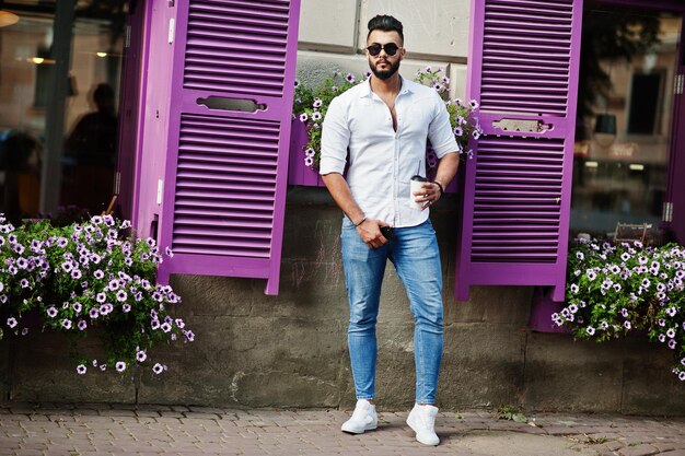 Modelo de hombre árabe alto con estilo en pantalones vaqueros de camisa blanca y gafas de sol posados en la calle de la ciudad Chico árabe atractivo de barba con una taza de café contra ventanas moradas