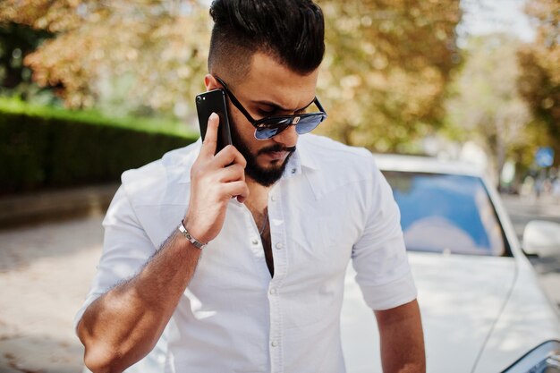 Modelo de hombre árabe alto con estilo en pantalones vaqueros de camisa blanca y gafas de sol posados en la calle de la ciudad Chico árabe rico y atractivo con barba contra un coche todoterreno blanco hablando por teléfono móvil