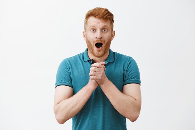 Modelo de hombre guapo e impresionado y emocionado alegre con cabello rojo y cerdas, con la mandíbula caída y jadeando mientras aprieta las palmas de las manos sobre el pecho y mira emocionado algo sorprendido y sorprendente