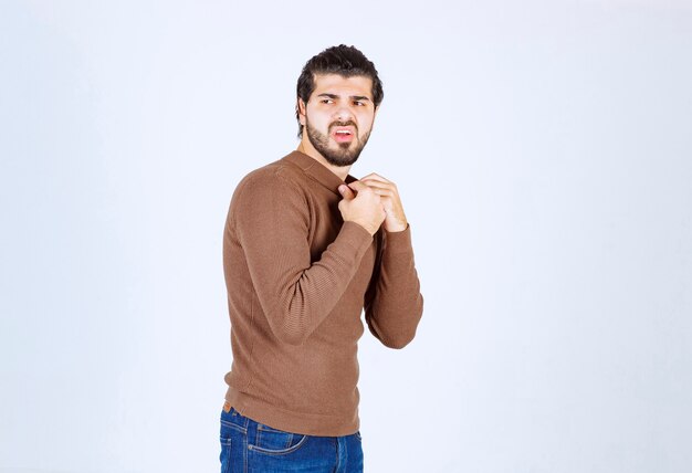 modelo de hombre guapo joven de pie en suéter marrón y posando sobre la pared blanca.