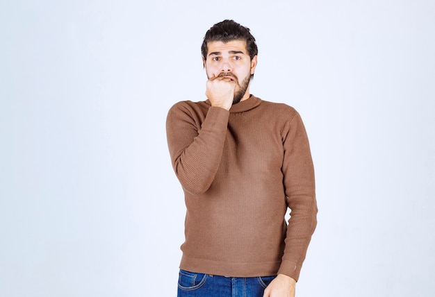 Un modelo de hombre joven de pie y mordiéndose las uñas sobre una pared blanca.