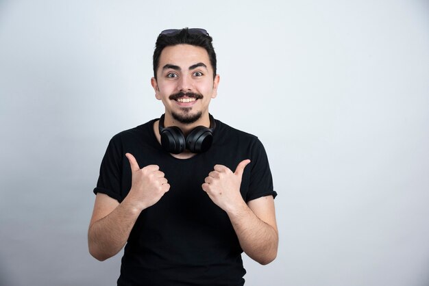 Modelo de hombre morena de pie en auriculares y mostrando los pulgares contra la pared blanca.