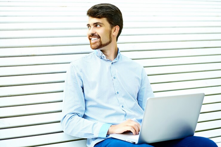 Foto gratuito modelo de hombre de negocios sonriente guapo joven sentado en el banco del parque usando laptop en tela casual hipster