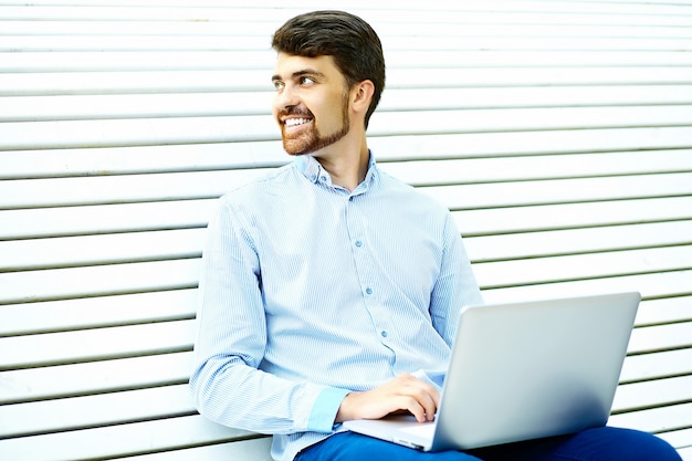 Modelo de hombre de negocios sonriente guapo joven sentado en el banco del parque usando laptop en tela casual hipster