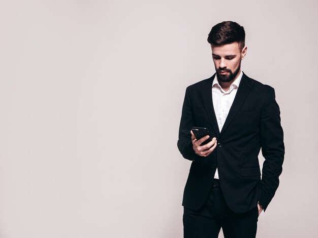 Modelo lambersexual de hipster elegante y seguro de sí mismo Hombre moderno sexy vestido con traje negro elegante Hombre de moda posando en el estudio Sosteniendo el teléfono inteligente Mirando la pantalla del teléfono celular Usando aplicaciones