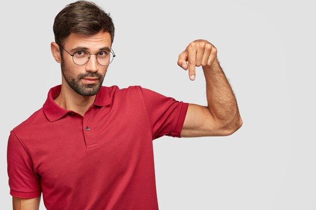 Modelo masculino sin afeitar seguro en gafas y camiseta roja, apunta hacia abajo