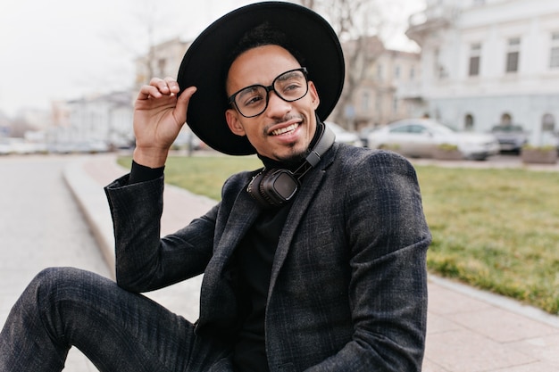 Modelo masculino africano de ensueño en traje de lana gris mirando a otro lado. Hombre joven extático con sombrero grande y gafas disfrutando de la sesión de fotos al aire libre.