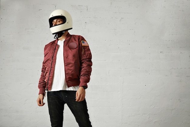 Modelo masculino atractivo joven en jeans negros, camiseta blanca lisa, chaqueta de bombardero de nylon burdeos y un casco de motocicleta blanco