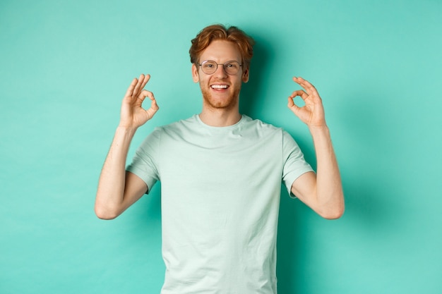 Modelo masculino atractivo con el pelo rojo, con gafas, mostrando OK firmar en aprobación y diciendo que sí, sonriendo satisfecho, de pie sobre fondo de menta.