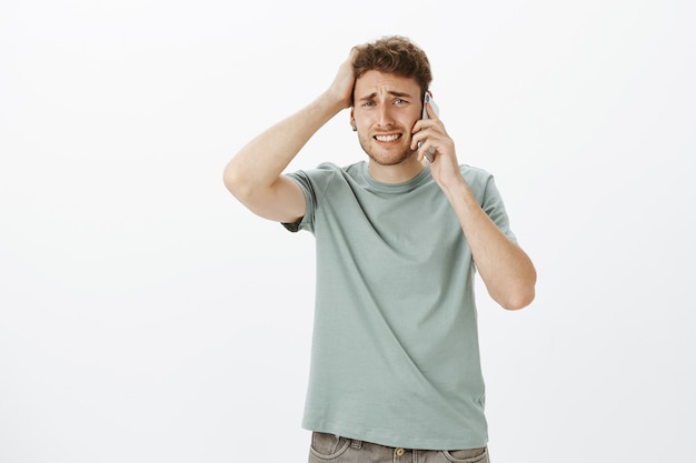 Modelo masculino guapo molesto y preocupado con cabello rubio, fruncir el ceño y apretar los dientes, tocar el cabello, sostener el teléfono inteligente cerca de la oreja, escuchar noticias terribles y estar triste