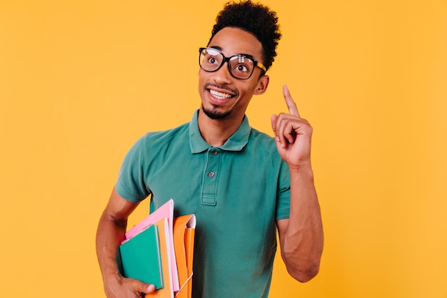 Modelo masculino inteligente con piel morena posando con libros. Retrato de interior de estudiante internacional dichoso en gafas de moda divirtiéndose.