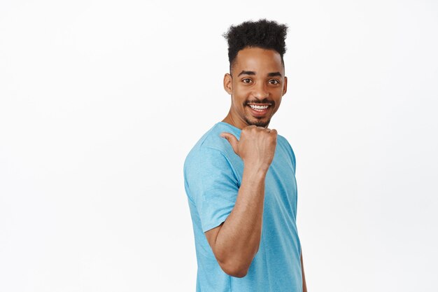 Modelo masculino joven guapo, chico afroamericano en camiseta que señala el dedo a la izquierda, detrás de su hombro, sonriendo y mirando feliz, invitar al evento en blanco
