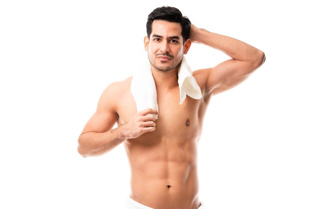 Modelo masculino latino con cuerpo musculoso sosteniendo una toalla alrededor de su cuello mientras está de pie contra el fondo blanco