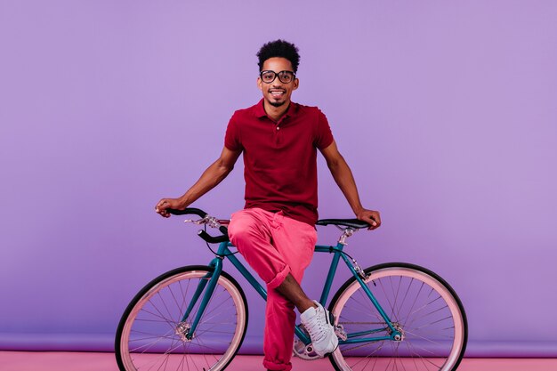 Modelo masculino negro alegre en pantalón rosa sentado en bicicleta. Filmación en interiores de niño africano riendo en vasos aislados.