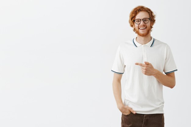 Modelo masculino en polo blanco apuntando a la izquierda con amplia sonrisa satisfecha y despreocupada de pie en gris