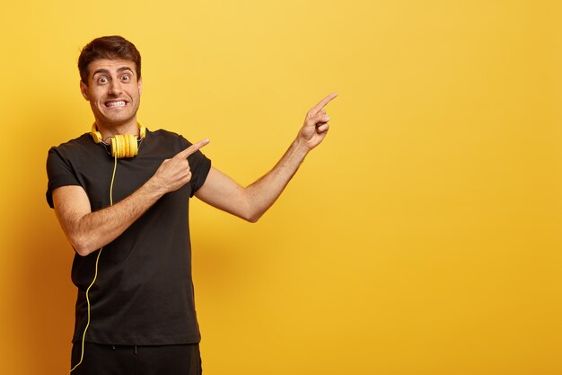 El modelo masculino positivo aprieta los dientes, señala un espacio en blanco para su texto o anuncio, usa auriculares en el cuello