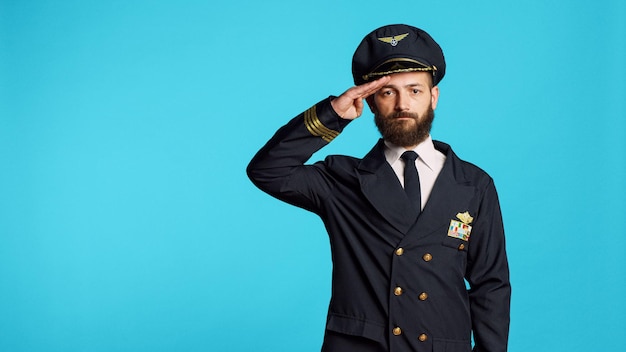 Modelo masculino con uniforme de piloto y sombrero en cámara, con ocupación profesional en la aerolínea. Joven trabajando como capitán y avión volador, siendo feliz y positivo en el estudio.