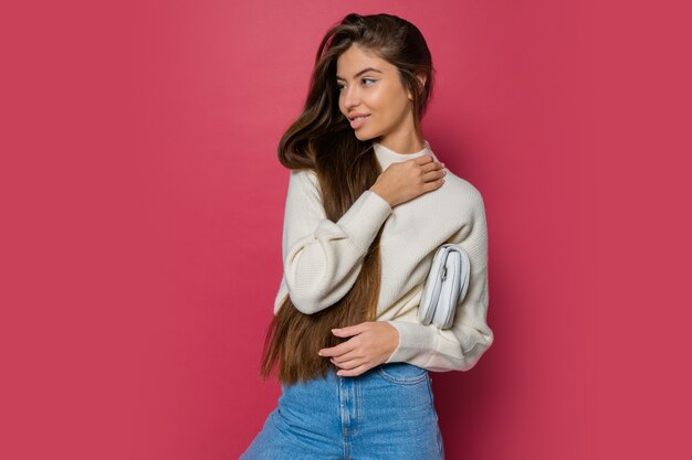 Modelo de moda en acogedor jersey blanco y jeans casuales posando sobre fondo rosa aislado. Sosteniendo el bolso de mano de cuero ecológico.