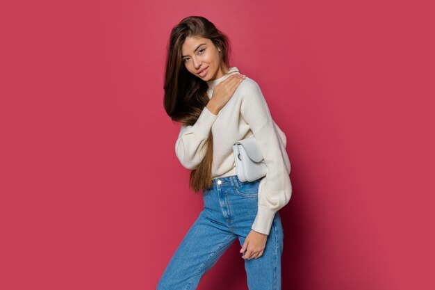 Modelo de moda en acogedor jersey blanco y jeans casuales posando sobre fondo rosa aislado. Sosteniendo el bolso de mano de cuero ecológico.