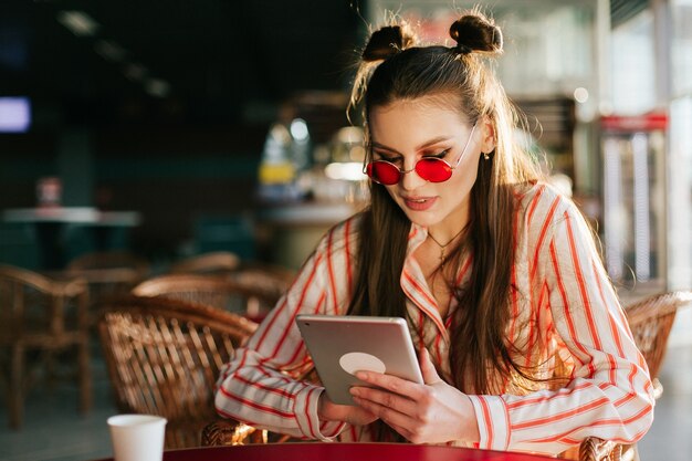 Modelo de moda bonito en gafas de sol rojos trabaja con su tableta sentado en el café