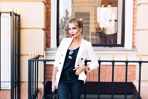 Modelo de moda en chaqueta blanca se apoya en la valla en la calle. Mantiene la mano en el bolsillo, mirando a un lado.