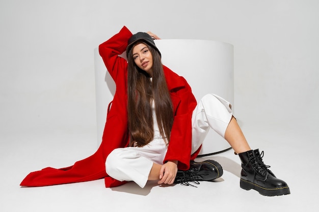 Modelo de moda en elegante sombrero, abrigo rojo y botas posando en la pared blanca en estudio