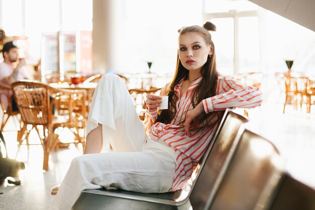 Modelo de moda joven se sienta con una taza de café en las sillas en el aeropuerto