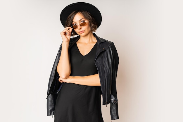 Modelo de moda con sombrero negro, vestido de noche y chaqueta de cuero posando en blanco.