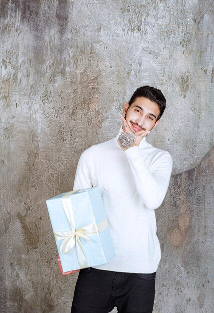 Modelo de moda en suéter blanco sosteniendo una caja de regalo azul envuelta con cinta blanca y parece pensativa o vacilante.