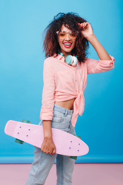 Modelo de mujer africana inspirada en gafas de sol amarillas divirtiéndose. Sonriente niña mulata con patineta posando en habitación con interior azul.