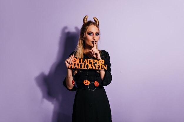 Modelo de mujer alegre en elegante vestido negro posando en halloween. adorable chica en traje de bruja de pie en la pared púrpura.