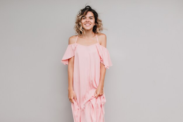 Foto gratuita modelo de mujer alegre con pelo rizado posando con una sonrisa sincera. foto interior de niña bronceada en vestido largo rosa aislado.