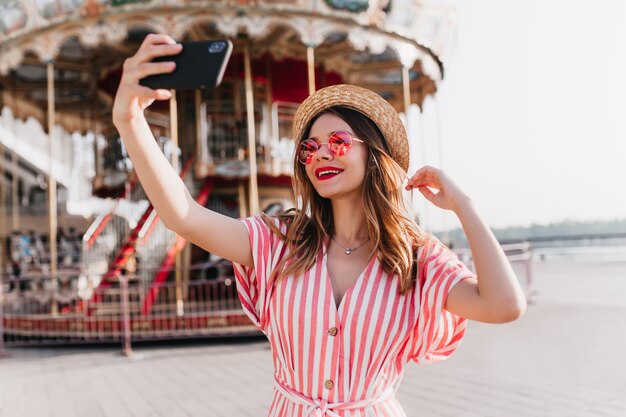 Modelo de mujer alegre en traje de rayas posando junto al carrusel con sombrero de paja. Tiro al aire libre de moda chica caucásica con smartphone para selfie en parque de atracciones.