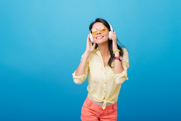 Modelo de mujer asiática inspirada en reloj de pulsera rosa y pulsera verde escuchando música. Foto interior de chica latina extática con gafas de sol naranjas tocando auriculares y sonriendo.