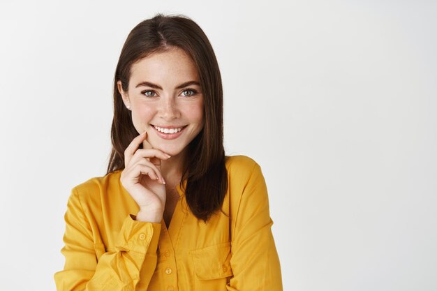 Modelo de mujer atractiva mirando con interés al frente, sonriendo y expresando felicidad, haciendo elección, de pie en la pared blanca.