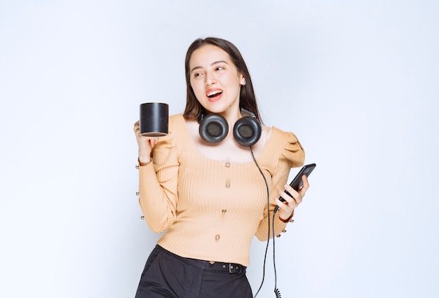 Un modelo de mujer atractiva sosteniendo una taza con teléfono móvil y auriculares.