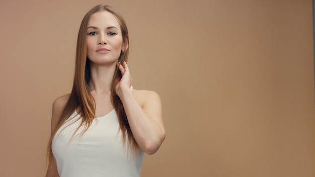 Modelo de mujer de belleza en estudio sobre fondo beige toca su cabello rubio recto y mira a la cámara