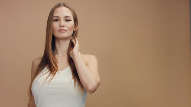 Modelo de mujer de belleza en estudio sobre fondo beige toca su cabello rubio recto y mira a la cámara