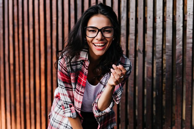 Modelo de mujer bronceada de ensueño posando emocionalmente en la pared de madera. Retrato al aire libre de una encantadora niña lleva gafas.