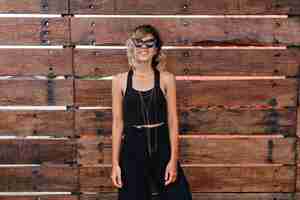 Foto gratuita modelo de mujer caucásica despreocupada en ropa negra de pie junto a la pared de madera. relajada joven con cabello rubio disfrutando de la sesión de fotos.
