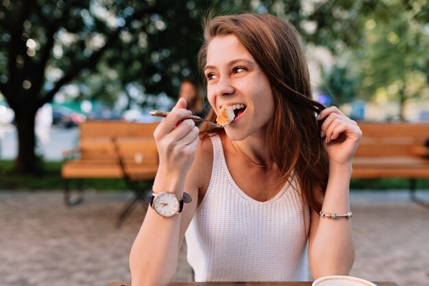 Modelo de mujer caucásica sonriente comiendo cupcake de lujo en la cafetería de verano al aire libre