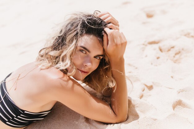 Modelo de mujer complacida con cabello oscuro posando mientras está acostado en la arena. Retrato al aire libre de una impresionante mujer alegre en traje de baño a rayas.