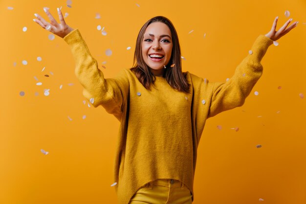 Modelo de mujer emocionada en suéter amarillo suave de pie bajo confeti. Retrato de mujer alegre con cabello lacio oscuro.