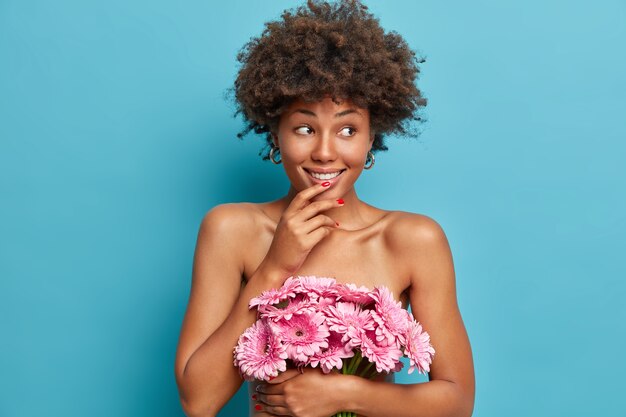 Modelo de mujer feliz sensual con cuerpo desnudo sano, sostiene ramo de flores de gerberas rosadas, mira con expresión alegre de ensueño a un lado, se encuentra