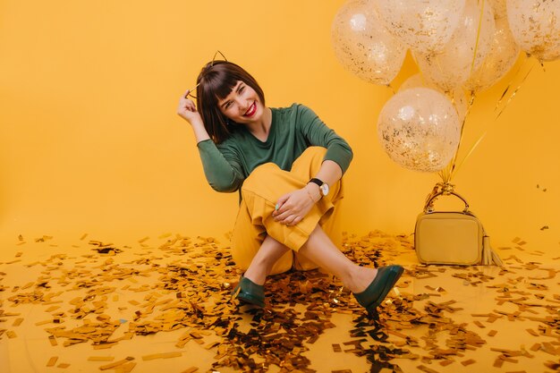 Modelo de mujer feliz sentada en el suelo cubierto de confeti y riendo. Encantadora chica caucásica posando con globos en su cumpleaños.