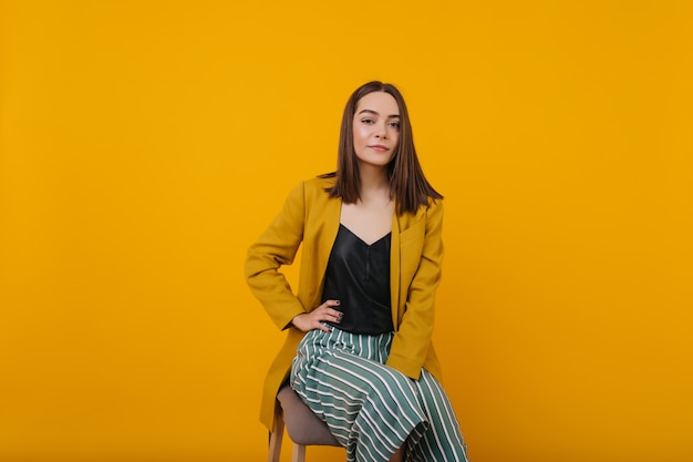 Modelo de mujer glamorosa con pelo lacio sentado en una silla. Retrato de interior de mujer joven impresionante en chaqueta amarilla de moda.