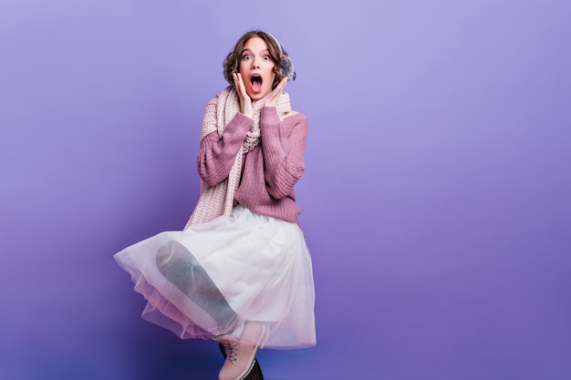 modelo de mujer guapa que expresa asombro sincero. Retrato de interior de una chica encantadora en falda blanca y bufanda posando en la pared púrpura.