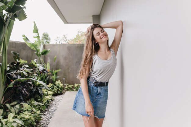 Modelo de mujer inspirada con un cabello precioso posando al aire libre cerca de las plantas. Retrato de mujer joven bien formada en falda de mezclilla de pie junto a la pared blanca con sonrisa romántica.